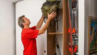 Man i röd tröja ställer upp en ryggsäck i en nybyggd garderob. Bredvid står skidor och en dammsugare.