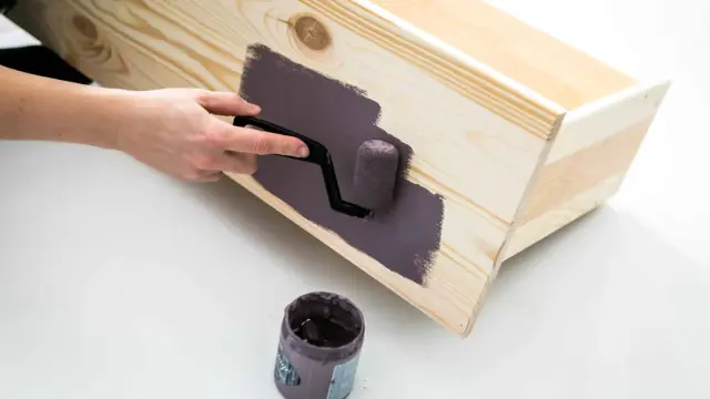 En hand använder en liten roller för att måla en träkista med mörklila färg, vilket ger en jämn yta utan synliga penseldrag.